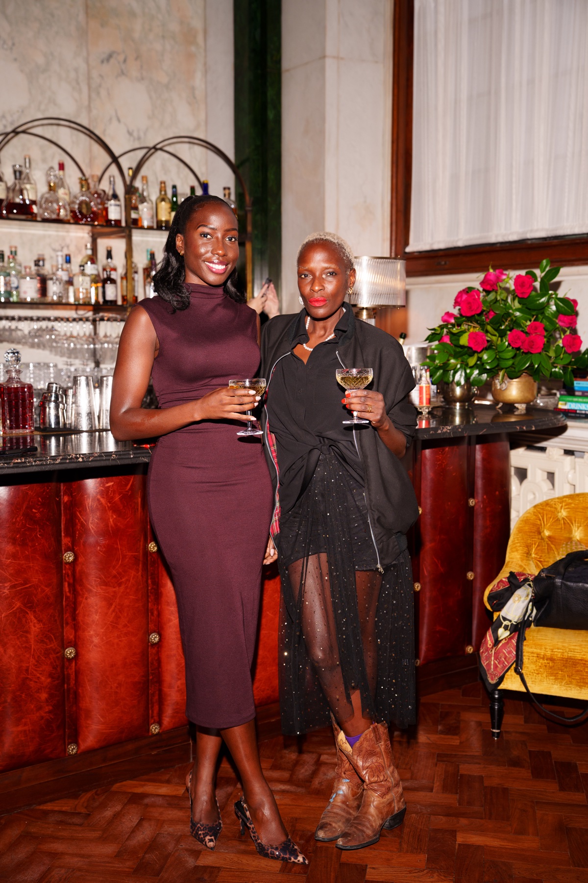 Two women holding cocktails beside a bar decorated with red roses