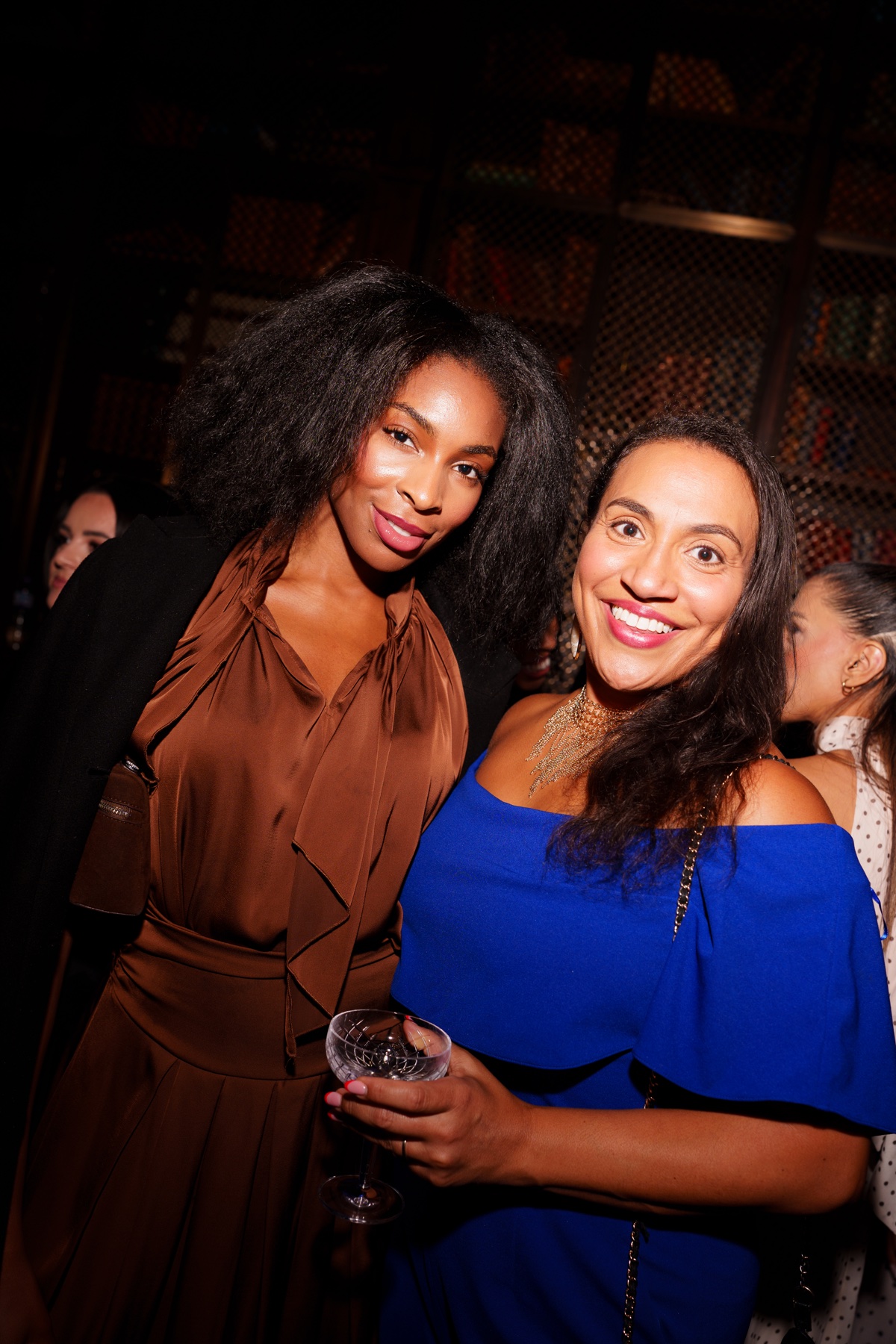 Two women in brown and blue outfits posing together with a cocktail