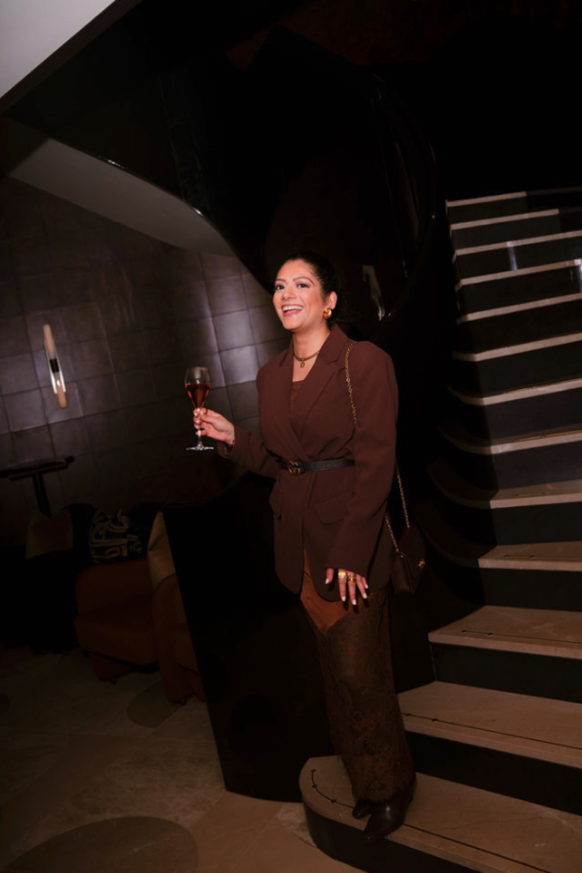 An elegantly dressed woman smiling with a glass beside a staircase