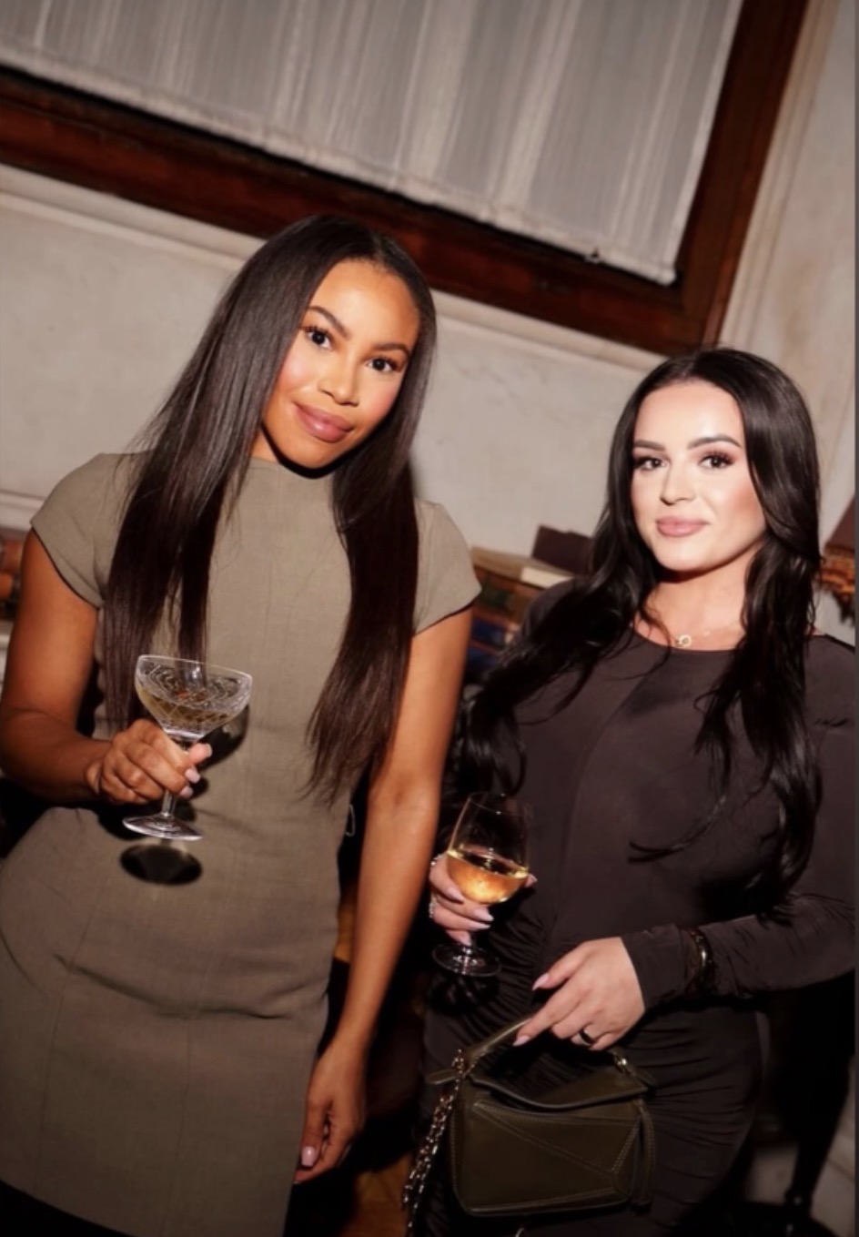 Two women in olive and charcoal dresses holding drinks together